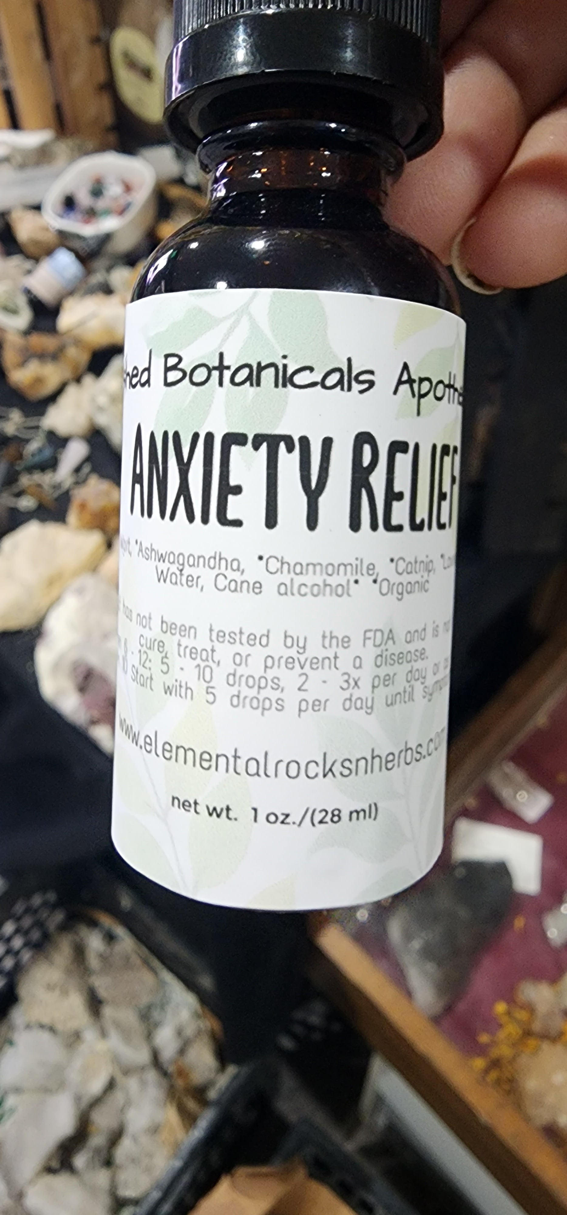 A hand holds a small bottle labeled "Anxiety Relief Tincture" from "Crushed Botanicals Apothecary." The supplement, designed to improve mood and provide stress relief, contains ashwagandha, chamomile, and other ingredients. The label states it is not FDA-approved for curing diseases and provides dosage instructions.