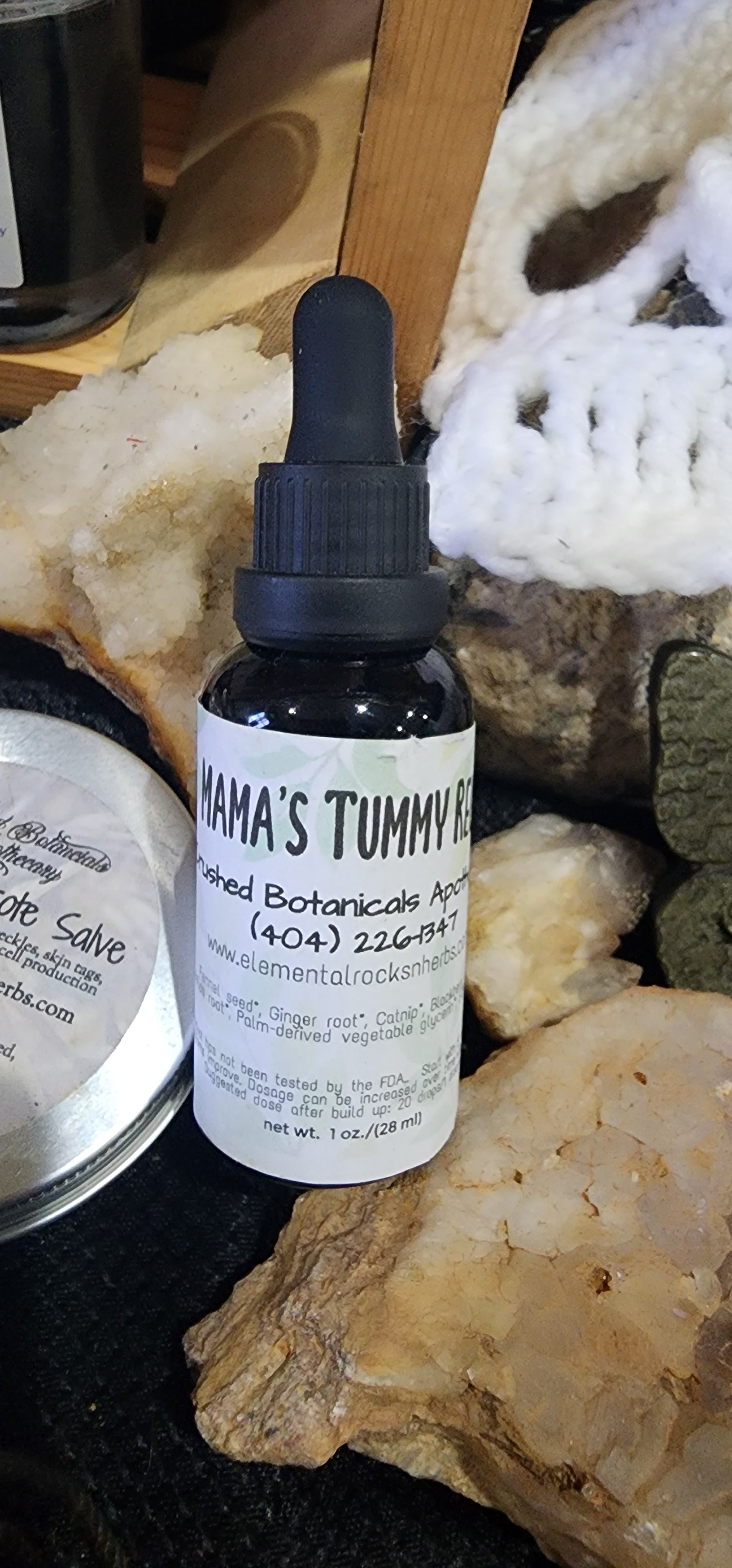 A dropper bottle labeled "Mama's Tummy Tonic Extract (Pregnancy Safe) - Morning Sickness" from "Crushed Botanicals Apothecary" sits among various natural items, including stones and a crocheted piece. Promising pregnant tummy relief, the bottle has a black dropper cap and a white label with contact information and ingredient details.